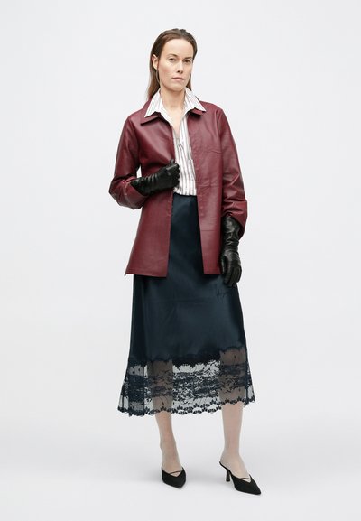 Γυναίκα που φοράει μπουφάν από δέρμα μπορντό, μαύρα δερμάτινα γάντια, πουκάμισο με ρίγες, μαύρη midi φούστα με δαντέλα στο τελείωμα και μαύρες γόβες σε λευκό φόντο.
