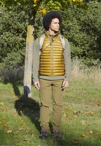 Brown puffer vest over a green long-sleeve shirt, paired with green pants and brown hiking boots. Standing in a natural setting.