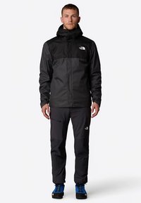 Chaqueta impermeable negra con capucha, con una sección superior gris contrastante y logotipo. Combinada con pantalones negros y zapatos de senderismo azules.