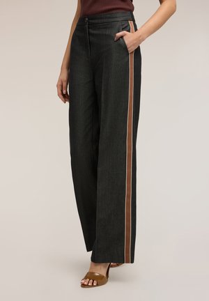 Žena v tmavých kalhotách s širokými nohavicemi a hnědými pruhy po stranách, má ruku v kapse, kombinuje je s hnědými sandály na podpatku a bezrukávovým topem.