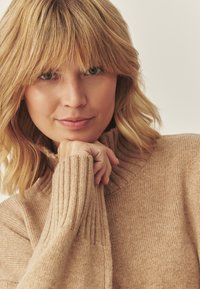 Maglione beige con collo alto, texture a coste, maniche aderenti e tessuto morbido. La modella poggia il mento sulla mano.