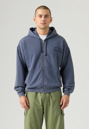 Mann in blauer Reißverschluss-Hoodie mit aufgesetzter Kapuze und grüner Cargo-Shorts, der vor einem einfarbigen hellen Hintergrund steht.