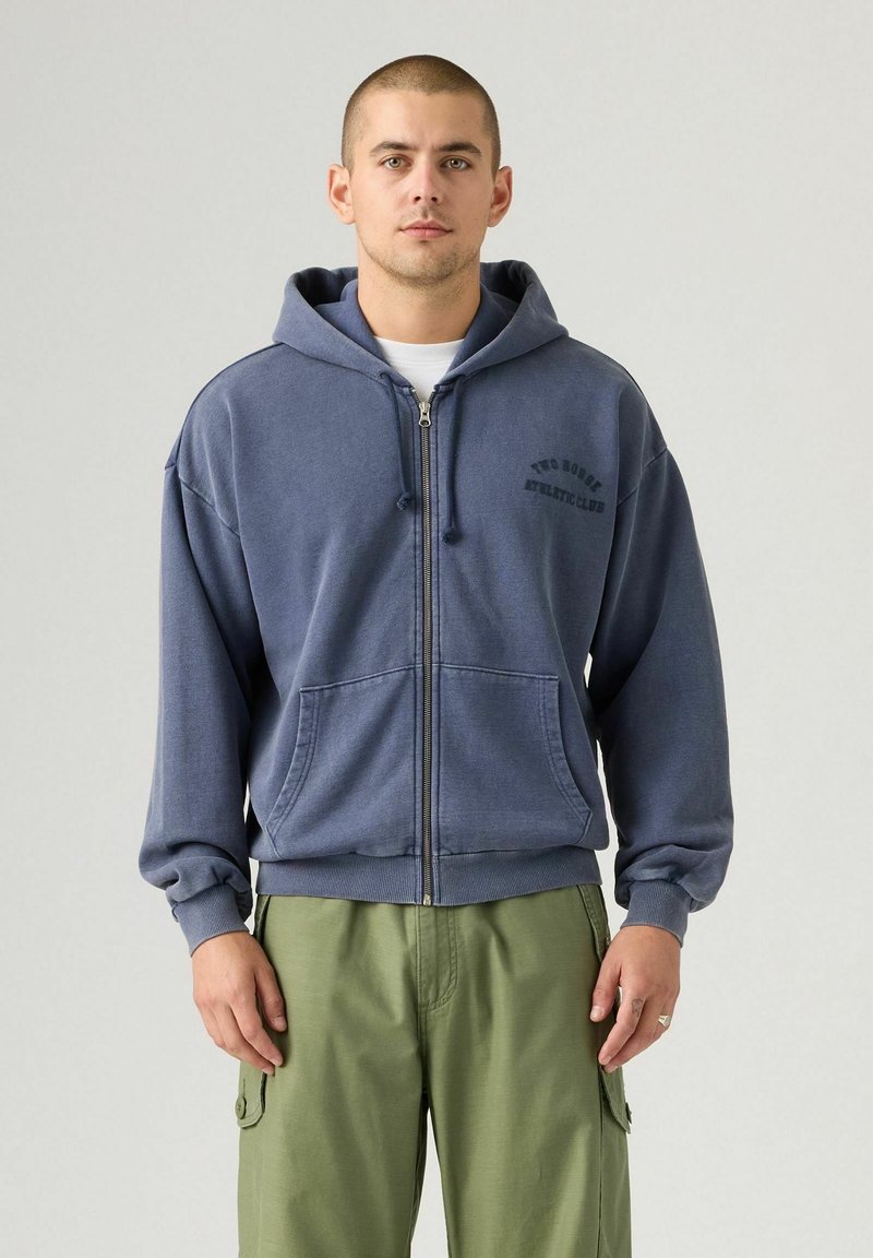 Hombre vestido con una sudadera azul con cremallera y capucha puesta, y pantalones cargo verdes, de pie contra un fondo claro y sencillo.