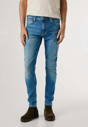 Mężczyzna w obcisłych niebieskich dżinsach z wyblakłym efektem i ciemnobrązowych zamszowych butach typu Chelsea, stojący na jednolitym tle.