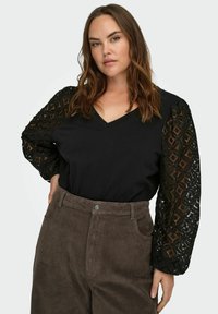 Czarna bluzka z dekoltem w serek i koronką, z bufkami na rękawach; skomplikowany wzór czarnej koronki. W zestawie z brązowymi sztruksowymi spodniami z przednimi kieszeniami i zapięciem na guziki.