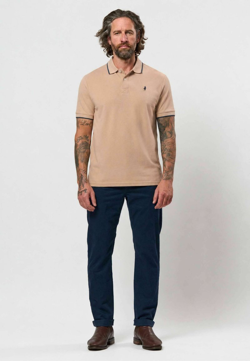 Homme aux cheveux longs et tatouages se tient face à l'avant, portant un polo beige, un pantalon bleu marine et des bottes marron sur un fond uni.