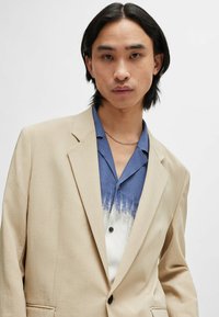 Beige Blazer mit Reversübergängen über einem blau-weißen Ombre-Hemd mit Knopfleiste. Der Blazer hat eine glatte Textur und einen einzelnen Knopfverschluss.