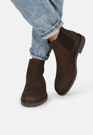Botas de ante marrón hasta el tobillo con paneles elásticos laterales, punteras redondeadas y suelas de goma, combinadas con unos vaqueros de mezclilla azul claro con vuelta.