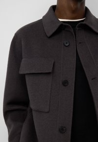 Ciemnoszara kurtka z wełny z kołnierzem, pojedynczą kieszenią na piersi i czarnymi guzikami. Miękka tekstura i dopasowany fason.