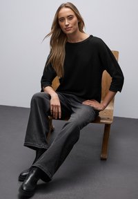 Černý texturovaný svetr, šedé rozšířené džíny a černé kotníkové boty. Sedící na dřevěné židli před světle šedým pozadím.