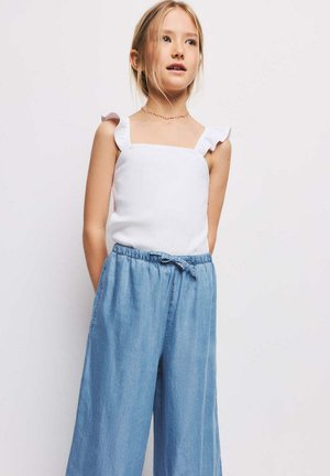 Jeune fille aux cheveux blonds portant un haut blanc sans manches avec des bretelles froncées et un pantalon ample en jean bleu à taille élastiquée avec cordon.