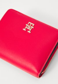 Portefeuille en cuir rouge avec un logo doré, fermeture éclair et texture lisse. Bords et coutures accentués visibles. Design compact.