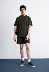 Olivengrøn t-shirt i bomuld, sorte mønstrede shorts, parret med grønne ruskindssneakers og hvide crew-sokker. Lille sort crossbody taske.