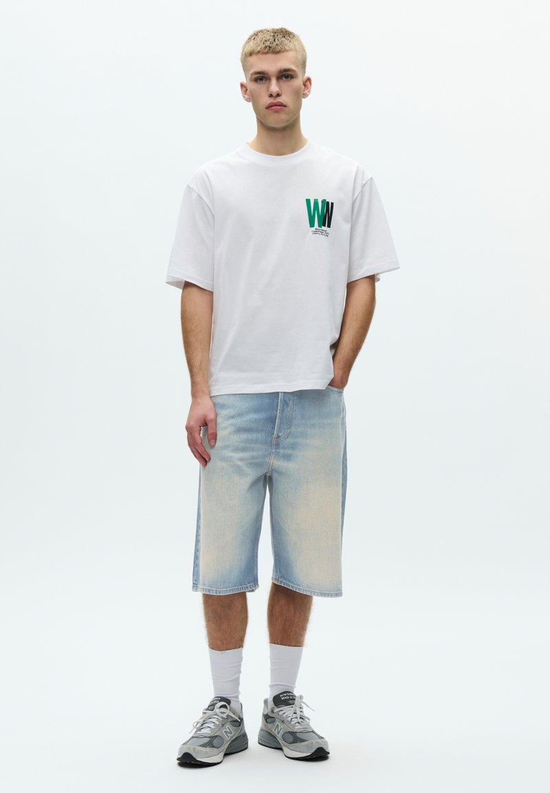 Weißes Baumwoll-T-Shirt mit grünem und schwarzem grafischem Logo, kombiniert mit hellblauen Jeans-Shorts. Das Modell trägt graue Sneakers und weiße Socken.