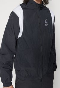 Černá lehká bunda s bílými panely na ramenou, na celou délku zip, elastickými manžetami, s logem Jumpman na hrudi a staženým pasem.