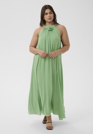 Femme portant une robe maxi dos nu vert clair avec un détail floral au niveau du décolleté et des sandales noires à lanières, debout devant un fond blanc.