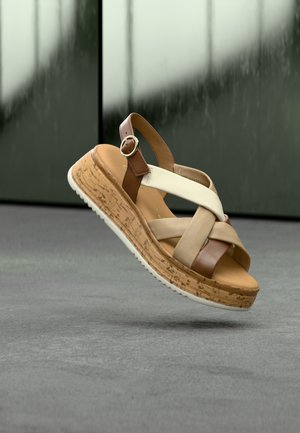Sandale compensée marron, beige et crème avec des brides croisées et une boucle, flottant au-dessus d'une surface grise avec un arrière-plan flou.