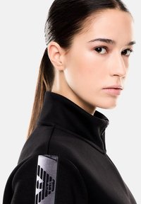 Sudadera negra de cuello alto con un parche en el hombro texturizado y un diseño gráfico en gris, que muestra un tejido suave y una silueta ajustada.
