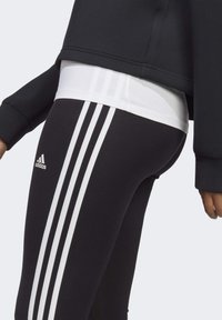 Sorte atletiske leggings med tre hvide striber langs siderne, parret med en cropped sort top og en hvid undertrøje.