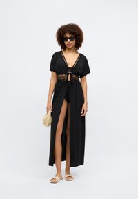 Μαύρο κάλυμμα παραλίας με σχέδιο κιμόνο, διακοσμημένο με πλέξη στο μέσο, ανοιχτό μπροστά και με ψηλή σχισμή. Φορεμένο πάνω από μαύρο μαγιό.