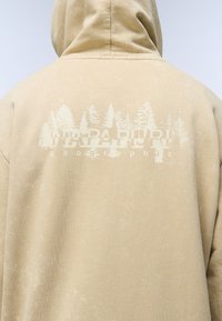 Sudadera con capucha beige con un acabado texturizado, con un gráfico de árboles y "NAPAPIJRI geographic" en blanco en la parte posterior.