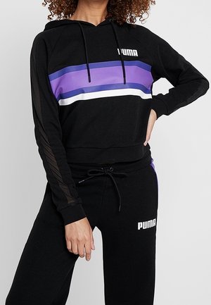 Woman wearing black Puma hoodie with purple and white stripes and matching black Puma sweatpants with drawstring waist.