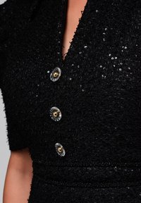 Robe noire texturée avec un décolleté en V, des manches courtes et trois boutons décoratifs avec des centres dorés et des entourages transparents.