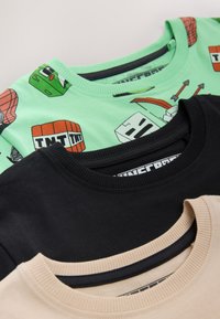 Tre crewneck T-shirts: en grøn med farverige grafikker, en sort og en beige. Hver har ribbet krave og blød bomuldstekstur.
