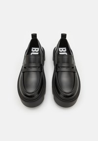 Une paire de mocassins en cuir noir avec des semelles épaisses et des détails de sangle, vue de dessus sur un fond blanc.