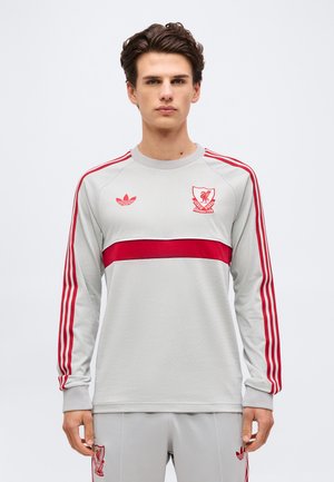 Νεαρός άνδρας με κοντά καστανά μαλλιά, ντυμένος με ανοιχτό γκρι φόρμα της Liverpool FC με κόκκινες ρίγες και λογότυπα, στέκεται μπροστά σε απλή φόντο.