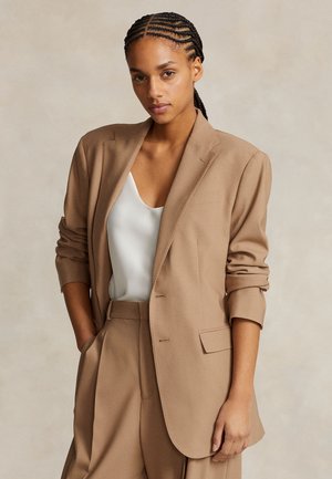 Frau mit geflochtenem Haar trägt einen beigen Blazer und eine Hose über einem weißen ärmellosen Oberteil und steht mit einer Hand in der Tasche vor einem schlichten Hintergrund.