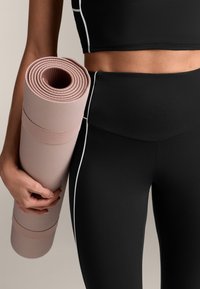 Črn športni crop top in visokovisentne leggins hlače z belimi poudarki, ki drži zvit rožnat teksturiran joga mat.