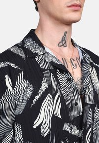 Camisa de manga corta en blanco y negro con un patrón de olas; presenta un cuello con solapa y cierre de botones. La tela tiene un aspecto texturizado y acanalado.