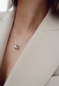 Sølv halskæde med en rund vedhæng med en glat overflade og en klar sten, båret over en lyse beige blazer.