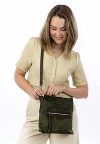 Young woman with shoulder-length hair wearing a beige buttoned top and pants, holding and opening a dark green crossbody bag.