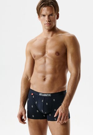 Mężczyzna bez koszulki, umięśniony, ubrany w czarne bokserki z białym napisem „ATLANTIC” na jednolitym jasnym tle.