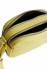 Žlutá kožená kabelka přes tělo s třemi zipovými přihrádkami a hladkým povrchem. Nastavitelný popruh připevněný kovovým hardwarem.