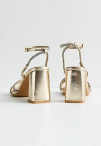 Gold heeled sandals with adjustable ankle straps. Features a wide block heel and embellishments of rhinestones along the straps. Smooth texture.