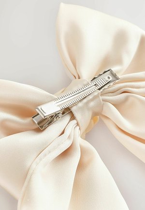 Cream-colored satin bow with a smooth texture, secured by a silver metal clip featuring a ridged design.