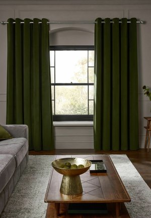 Wohnzimmer mit dunkelgrünen Vorhängen, die ein Fenster einrahmen, einem grauen Sofa, einem hölzernen Couchtisch mit goldener Schalen-Dekoration und einem hellen Teppich auf Holzboden.