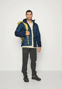 Navy insulated jacket, beige shirt, black pants, and black shoes. Model holds a yellow backpack, showcasing a casual outdoor look.
