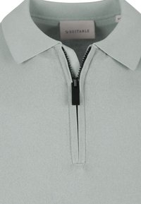Lichtgrijze polo met een korte rits, met een kraag en een gebreide textuur. Bevat een label met de tekst "Geschikt."