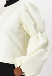 Blouse courte à manches bouffantes en satin jaune pâle avec un col rond et des poignets élastiques, présentant une texture lisse et nette.