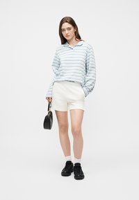 Νεαρή γυναίκα που φοράει μακρυμάνικο πουκάμισο με ανοιχτό μπλε ρίγες, λευκά σορτς, μαύρα αθλητικά παπούτσια, λευκές κάλτσες, κρατώντας μια μικρή μαύρη τσάντα, στέκεται μπροστά σε λευκό φόντο.