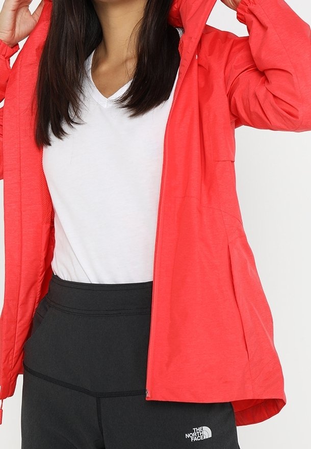 Veste légère rouge avec capuche, fermeture éclair à l'avant et poignets élastiques. Portée sur un tee-shirt blanc à col en V et un pantalon noir arborant un logo.