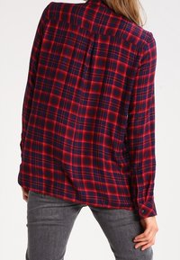 Chemise à carreaux rouge et bleu marine avec un col boutonné, des manches longues et une coupe décontractée. Présente un détail de empiècement au dos.