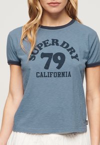 Niebieska koszulka z okrągłym dekoltem i krótkimi rękawami, z granatowym wykończeniem i odważnym nadrukiem z napisem "SUPERDRY 79 CALIFORNIA."