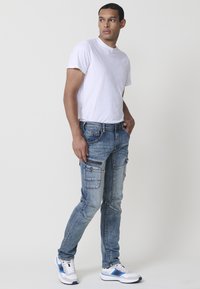Pantalones cargo de mezclilla en azul claro con múltiples bolsillos, corte ajustado, detalles descoloridos y costuras en contraste, combinados con una camiseta blanca y zapatillas.