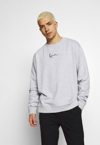 Grå sweatshirt med rund halsudskæring, blød tekstur og sort håndskrevet logo på brystet. Kombineret med sorte bukser.
