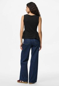 Crna majica bez rukava s peplum dizajnom na leđima, uparena s tamnoplavim širokim trapericama i smeškim cipelama s crvenim detaljima.